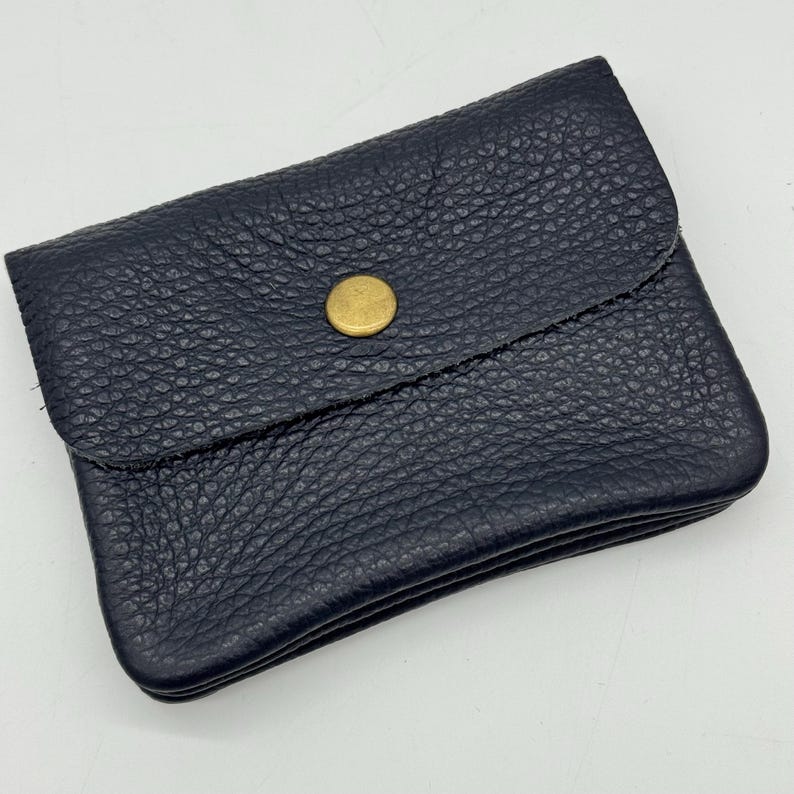 May include: A navy blue leather wallet with a textured surface. The wallet has a flap closure secured by a gold-colored button. The wallet is rectangular and appears to be folded.