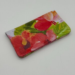 May include: A rectangular wallet with a floral design. The wallet features a vibrant pattern of red and pink flowers with green leaves against a light background. The wallet is closed and appears to be made of a smooth material.