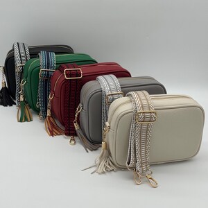 May include: A selection of rectangular crossbody bags in various colours, including black, green, burgundy, grey, and cream. Each bag features a patterned shoulder strap and a tassel. The bags are arranged in a row, showcasing their different hues.