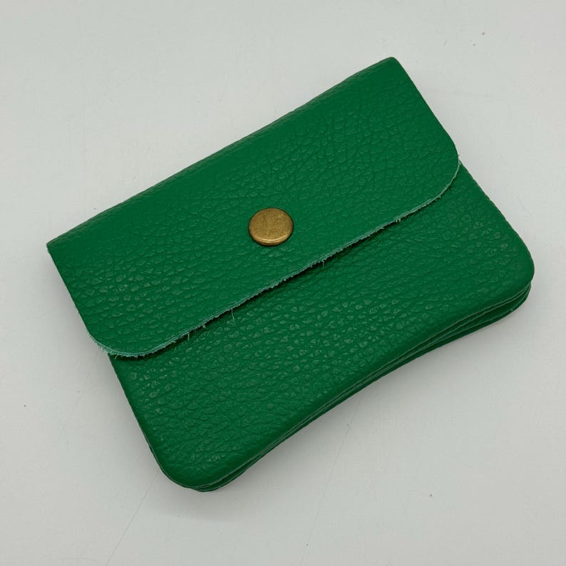 May include: A small, green leather wallet with a textured surface. The wallet has a flap closure secured by a gold-colored button. The wallet is rectangular and appears to be empty.