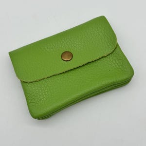 May include: A small, lime green leather wallet with a textured surface. The wallet has a flap closure secured by a round, antique brass-colored snap. The wallet is rectangular and appears to be empty.