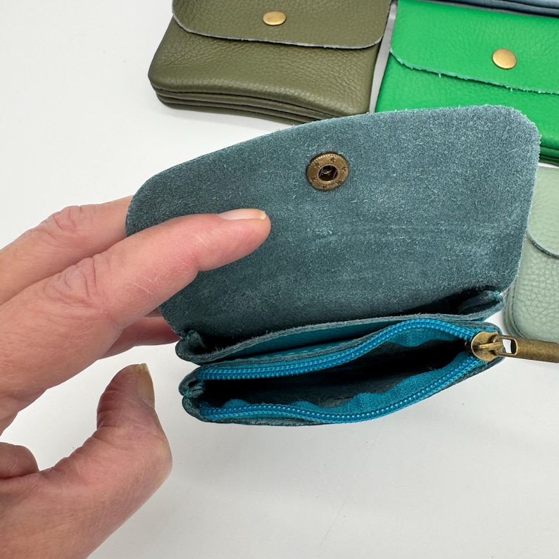 May include: Open teal leather coin purse with a brass snap closure and a teal zipper pocket. Other leather coin purses in green and olive are visible in the background.
