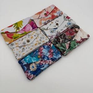 May include: A collection of seven floral print wallets in various colors. Each wallet features a flap closure with a brass-colored snap. The floral patterns include pink, yellow, blue, and red blooms on a white or dark background.