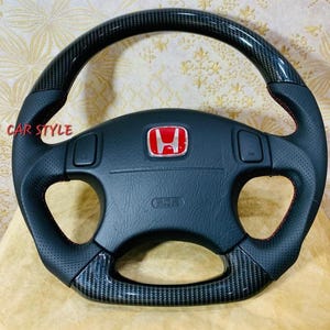 Può includere: Volante nero con dettagli in fibra di carbonio e cuciture rosse. Presenta il logo Honda in rosso e la scritta "CAR STYLE". Include pelle traforata e informazioni sull'airbag.