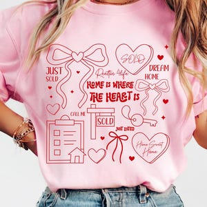 以下が含まれることがあります： ピンク色のTシャツ。赤いグラフィックデザインで、ハート、リボン、鍵、「Just Sold」、「Dream Home」、「Home is Where the Heart Is」の文字が描かれています。家とクリップボードも含まれています。