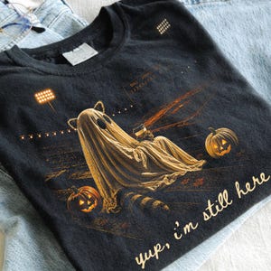 May include: A black t-shirt featuring a graphic of a ghost wearing cat ears and holding a coffee cup, sitting on a football field at night with two jack-o'-lanterns. The text "yup, i'm still here" is printed in a light tan script font at the bottom.