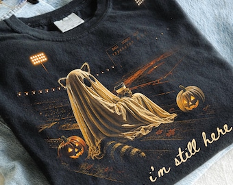 Może przedstawiać: Czarny t-shirt z grafiką ducha w kocich uszach, trzymającego kubek kawy, siedzącego nocą na boisku do futbolu z dwiema dyniami. Napis "yup, i'm still here" jest nadrukowany jasną czcionką w dolnej części koszulki.