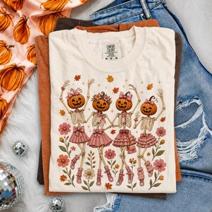 May include: A cream-colored short-sleeve t-shirt featuring a graphic of four skeletons with pumpkin heads wearing pink skirts and bows, surrounded by autumn leaves and flowers. The shirt has a Comfort Colors label at the inner neckline.