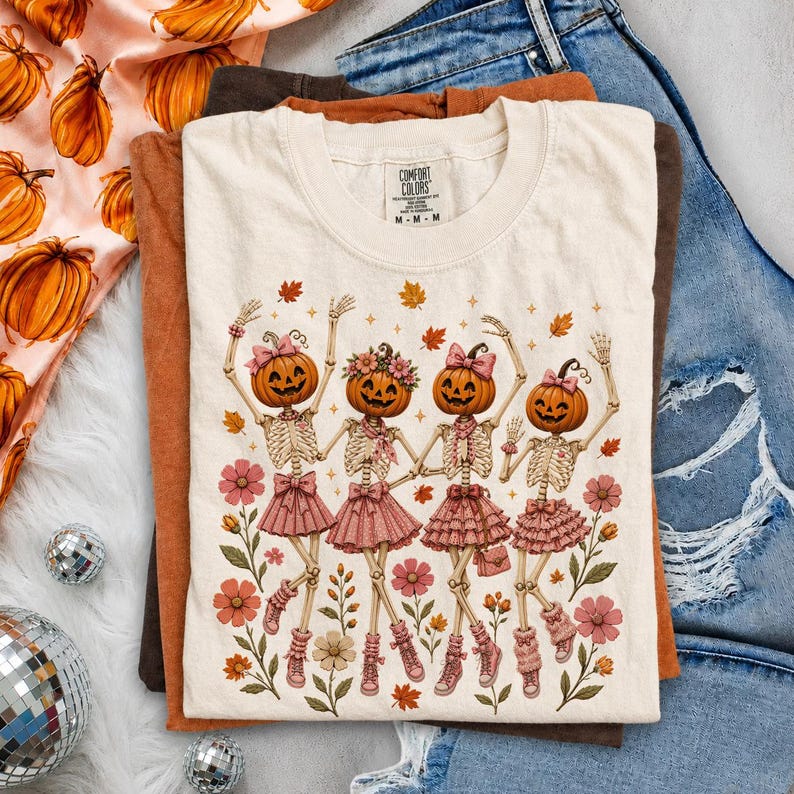 May include: A cream-colored short-sleeve t-shirt featuring a graphic of four skeletons with pumpkin heads wearing pink skirts and bows, surrounded by autumn leaves and flowers. The shirt has a Comfort Colors label at the inner neckline.