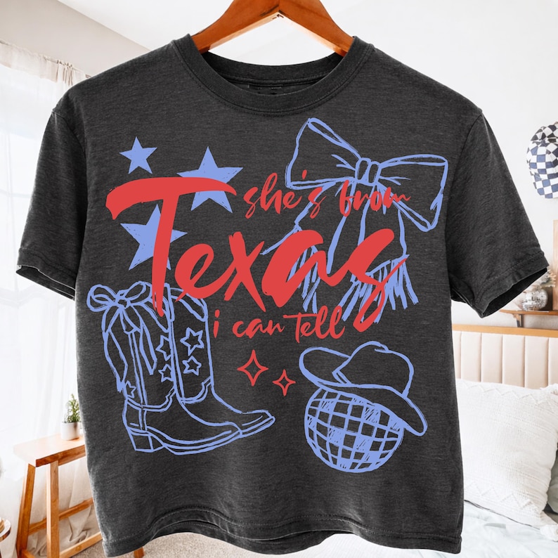 May include: Dark gray t-shirt with a Texas-themed design. The design includes the words "Texas" in red and blue, along with stars, a bow, cowboy boots, and a hat. The text "she's from Texas I can tell" is also included.
