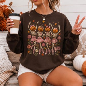 May include: A dark brown crewneck sweatshirt featuring a graphic print of four skeletons with pumpkin heads wearing pink skirts and bows. The skeletons are surrounded by small orange and pink flowers and autumn leaves, creating a festive seasonal design.