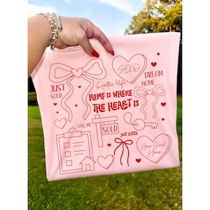 May include: A light pink t-shirt with red graphics related to real estate. The design includes hearts, bows, a house, a key, and the words "Just Sold," "Dream Home," and "Home Sweet Home." The phrase "Home is where the heart is" is also featured.