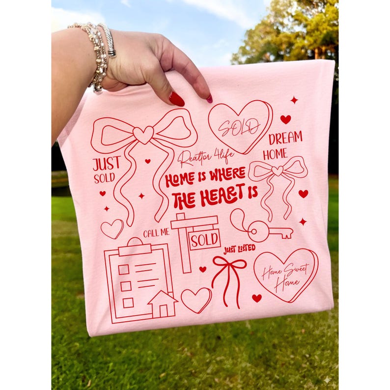 May include: A light pink t-shirt with red graphics related to real estate. The design includes hearts, bows, a house, a key, and the words "Just Sold," "Dream Home," and "Home Sweet Home." The phrase "Home is where the heart is" is also featured.