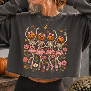 May include: A charcoal gray long-sleeve sweatshirt featuring a graphic print of four skeletons with pumpkin heads. Each skeleton wears a pink ruffled skirt, pink hair bows, and pink sneakers, surrounded by orange autumn leaves and pink flowers.