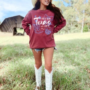 May include: Maroon sweatshirt with the text "Texas" and the phrase "she's from I can tell" in white script. The design includes a cowboy boot, a bow, a star, a disco ball, and a cowboy hat in blue. The person is wearing denim shorts and white cowboy boots.