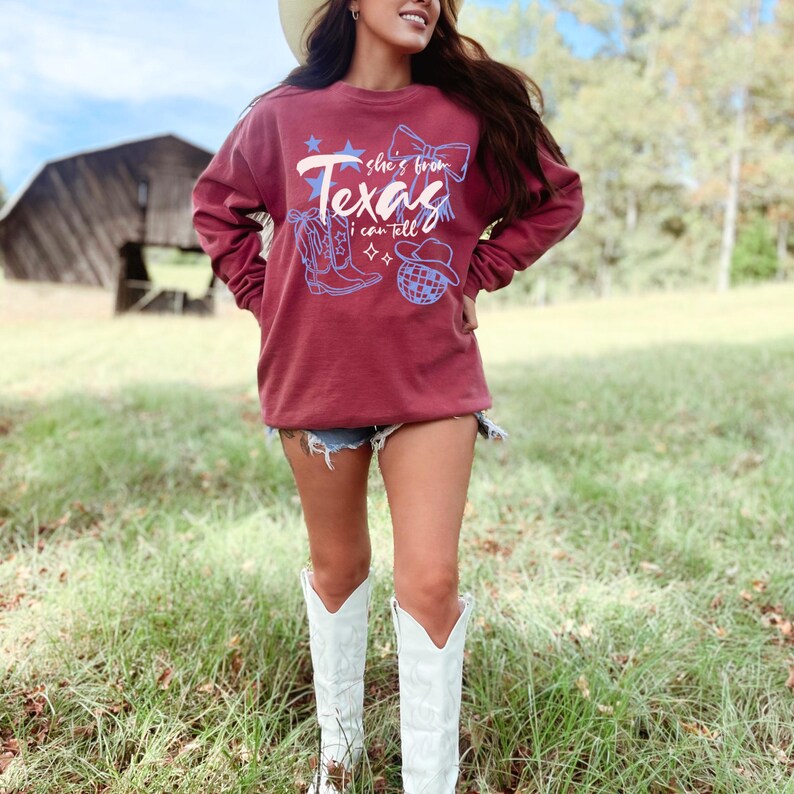 May include: Maroon sweatshirt with the text "Texas" and the phrase "she's from I can tell" in white script. The design includes a cowboy boot, a bow, a star, a disco ball, and a cowboy hat in blue. The person is wearing denim shorts and white cowboy boots.