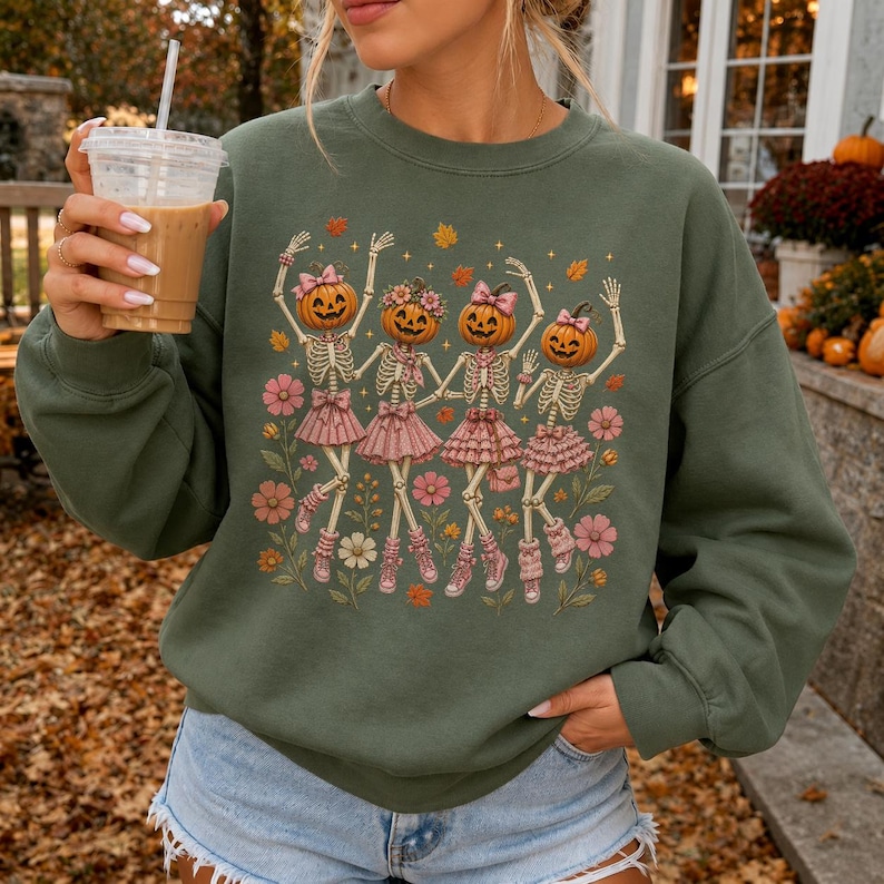 May include: An olive green crewneck sweatshirt featuring a graphic print of four skeletons with pumpkin heads wearing pink ruffled skirts and sneakers, surrounded by pink flowers and orange autumn leaves on a long-sleeved cotton garment.
