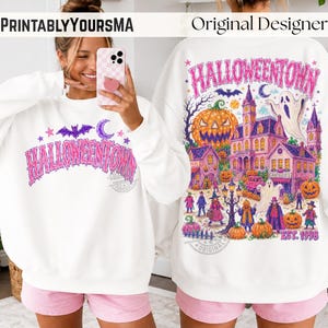 May include: A white long-sleeve sweatshirt featuring a pink and purple graphic print. The front displays the text HALLOWEENTOWN. The back features a detailed illustration of a haunted house, pumpkins, ghosts, and the text HALLOWEENTOWN EST. 1998.