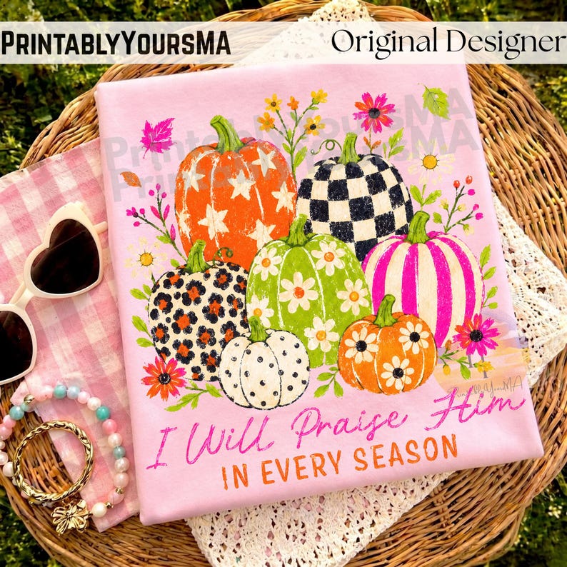 May include: A light pink t-shirt featuring a graphic of six pumpkins with various patterns including stars, leopard print, checkers, and stripes, surrounded by flowers. The text reads, "I Will Praise Him IN EVERY SEASON" in pink and orange.