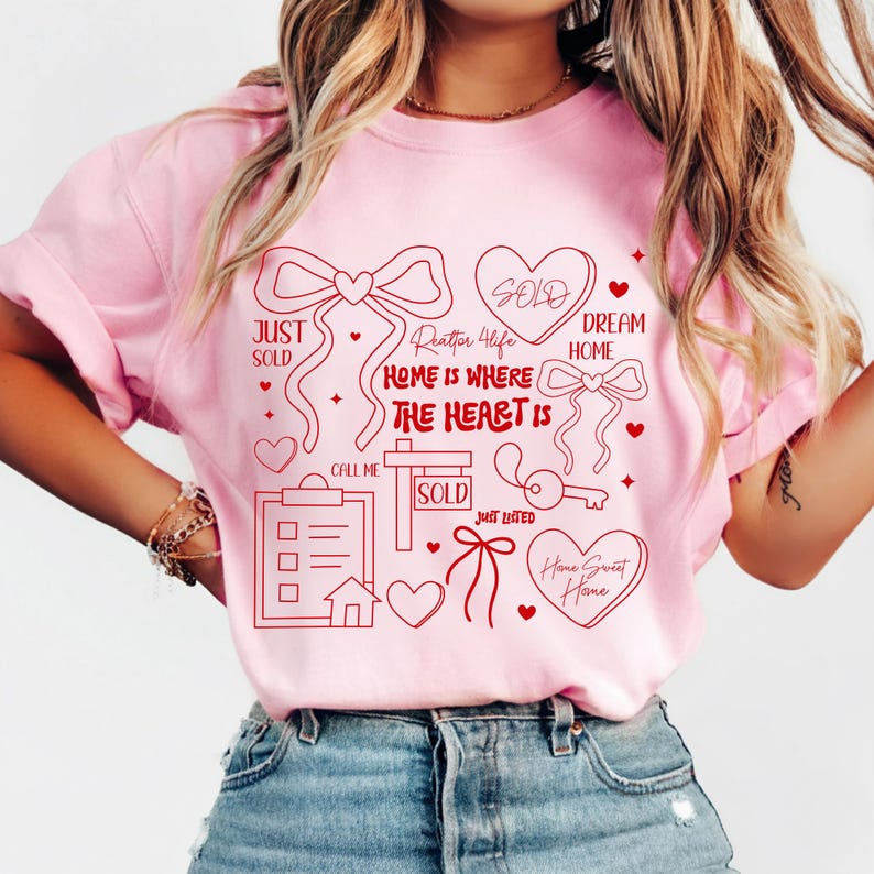 May include: Pink t-shirt with a red graphic design featuring hearts, bows, a key, and the words "Just Sold," "Dream Home," and "Home is Where the Heart Is." The design also includes a house and a clipboard.