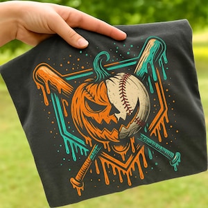 Puede incluir: Camiseta gris oscuro con un diseño de Halloween. El gráfico combina una calabaza de Halloween y una pelota de béisbol, con detalles goteantes en naranja y verde azulado. Dos bates de béisbol se cruzan detrás del diseño.