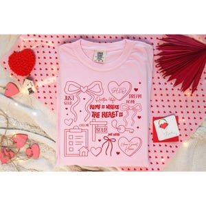 May include: A light pink t-shirt with red graphics and text. The design includes hearts, bows, a key, and the words "Just Sold," "Home is Where The Heart Is," and "Dream Home." The shirt is a Comfort Colors brand.