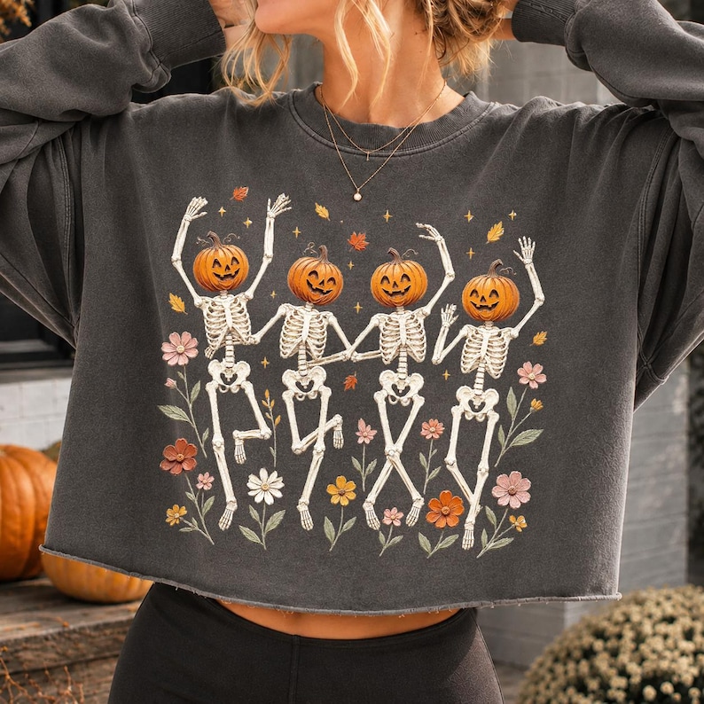 K&ouml;nnte beinhalten: Ein anthrazitfarbenes Langarm-Sweatshirt mit einem Grafikdruck von vier Skeletten mit K&uuml;rbisk&ouml;pfen, die zwischen bunten Blumen und fallenden Bl&auml;ttern tanzen. Das Kleidungsst&uuml;ck hat einen Rundhalsausschnitt und einen rohen, unges&auml;umten Saum.