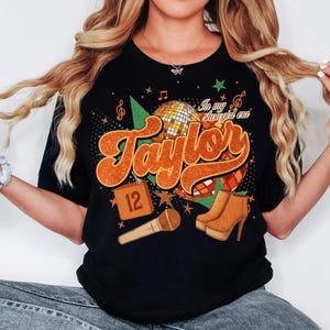 K&ouml;nnte beinhalten: Schwarzes T-Shirt mit einem Retro-Design mit dem Namen "Taylor" in Orange, einer Discokugel und einem Mikrofon. Das Design enth&auml;lt den Text "In my shougirl era" und die Nummer "12". Braune High-Heel-Stiefel sind ebenfalls abgebildet.