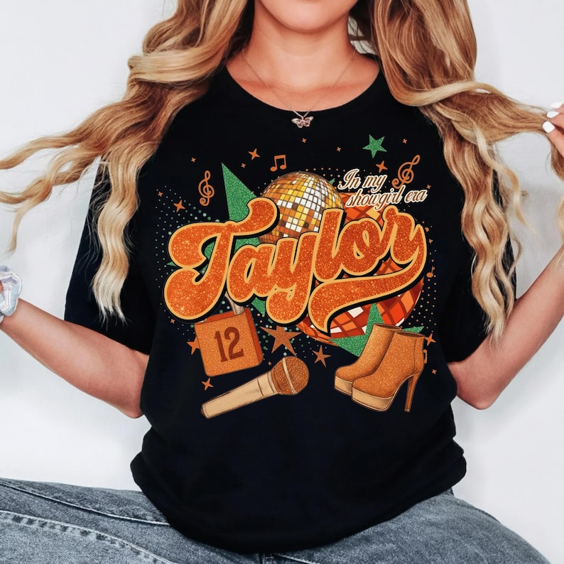K&ouml;nnte beinhalten: Schwarzes T-Shirt mit einem Retro-Design mit dem Namen "Taylor" in Orange, einer Discokugel und einem Mikrofon. Das Design enth&auml;lt den Text "In my shougirl era" und die Nummer "12". Braune High-Heel-Stiefel sind ebenfalls abgebildet.