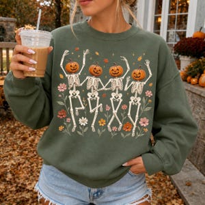K&ouml;nnte beinhalten: Ein olivgr&uuml;ner Rundhals-Pullover mit einem Grafikdruck von vier Skeletten mit K&uuml;rbisk&ouml;pfen, die zwischen bunten Herbstblumen und fallenden Bl&auml;ttern tanzen. Die Skelette sind wei&szlig; und zeigen detaillierte Rippenk&auml;fige und Gliedma&szlig;en.