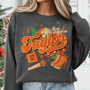 K&ouml;nnte beinhalten: Dunkelgraues Sweatshirt mit einem orangefarbenen Aufdruck mit dem Schriftzug "Taylor", einer Discokugel, einem Mikrofon und High-Heel-Stiefeln. Der Text "In my shougirl era" ist ebenfalls enthalten. Eine 12 ist ebenfalls abgebildet.
