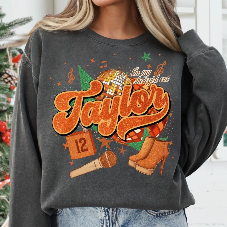 K&ouml;nnte beinhalten: Dunkelgraues Sweatshirt mit einem orangefarbenen Aufdruck mit dem Schriftzug "Taylor", einer Discokugel, einem Mikrofon und High-Heel-Stiefeln. Der Text "In my shougirl era" ist ebenfalls enthalten. Eine 12 ist ebenfalls abgebildet.