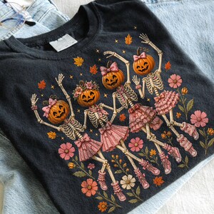 May include: A black t-shirt featuring a graphic of four skeletons with pumpkin heads, wearing pink ruffled skirts, bows, and sneakers. The skeletons are surrounded by orange autumn leaves and pink flowers, creating a festive seasonal design.