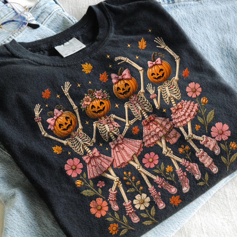 May include: A black t-shirt featuring a graphic of four skeletons with pumpkin heads, wearing pink ruffled skirts, bows, and sneakers. The skeletons are surrounded by orange autumn leaves and pink flowers, creating a festive seasonal design.