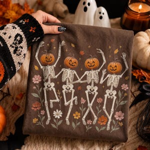 K&ouml;nnte beinhalten: Ein dunkelbraunes T-Shirt mit einem Grafikdruck von vier Skeletten mit orangefarbenen K&uuml;rbisk&ouml;pfen, die in einer Reihe tanzen. Die Skelette sind von kleinen bunten Blumen und Herbstbl&auml;ttern umgeben, mit winzigen goldenen Sternen auf dem Stoff.