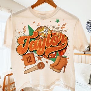 K&ouml;nnte beinhalten: Cremefarbenes T-Shirt mit Retro-Design mit dem Namen "Taylor" in orangefarbenen Glitzerbuchstaben. Das Design umfasst eine Discokugel, ein Mikrofon, Stiefel und den Text "In my showgirl era".