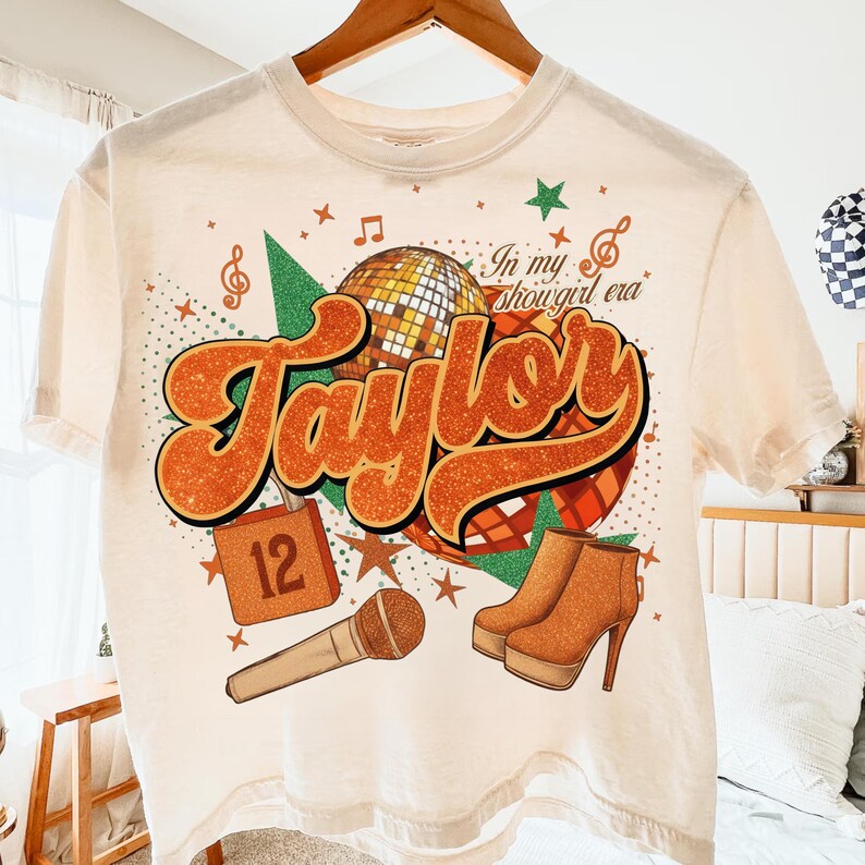K&ouml;nnte beinhalten: Cremefarbenes T-Shirt mit Retro-Design mit dem Namen "Taylor" in orangefarbenen Glitzerbuchstaben. Das Design umfasst eine Discokugel, ein Mikrofon, Stiefel und den Text "In my showgirl era".