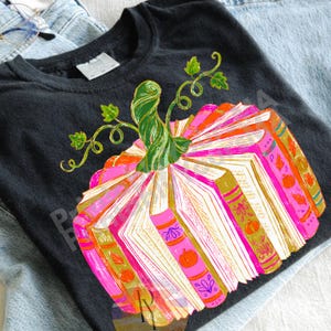 May include: A black t-shirt featuring a graphic of a pumpkin constructed from open books. The books have bright pink and orange spines with gold accents and small pumpkin illustrations. A green, swirling pumpkin stem sits on top.