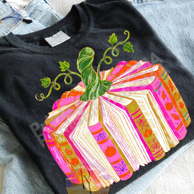May include: A black t-shirt featuring a graphic of a pumpkin constructed from open books. The books have bright pink and orange spines with gold accents and small pumpkin illustrations. A green, swirling pumpkin stem sits on top.