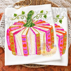 May include: A white t-shirt featuring a graphic illustration of a pumpkin constructed from stacked books. The books are coloured in shades of pink, orange, and gold, decorated with small pumpkin and leaf motifs. A green stem with curling vines sits on top.