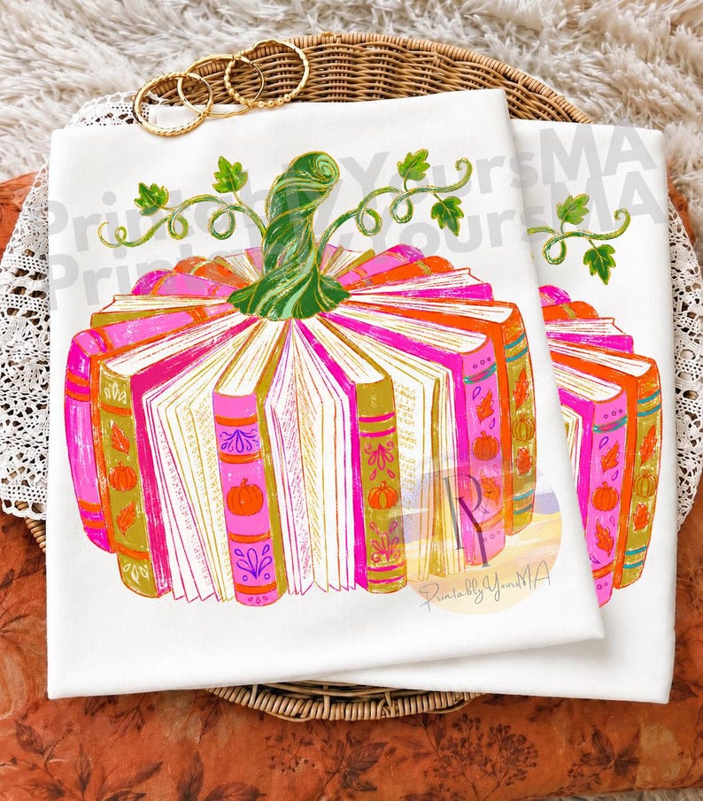 May include: A white t-shirt featuring a graphic illustration of a pumpkin constructed from stacked books. The books are coloured in shades of pink, orange, and gold, decorated with small pumpkin and leaf motifs. A green stem with curling vines sits on top.