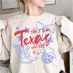 May include: A beige sweatshirt with a Texas-themed graphic. The design includes the words "she's from Texas I can tell" in red and blue, along with illustrations of stars, cowboy boots, a bow, and a disco ball.