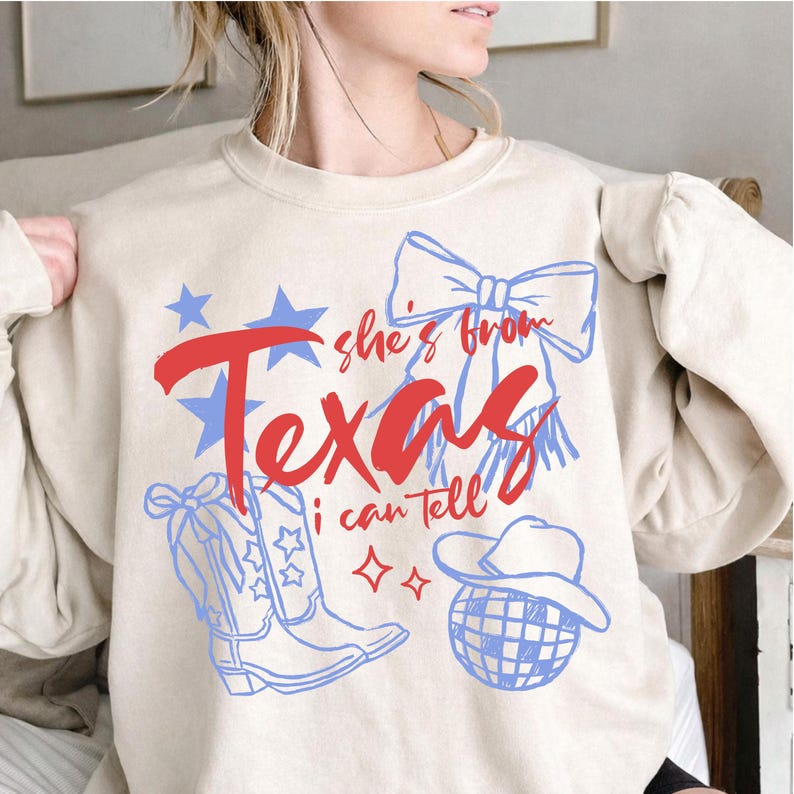 May include: A beige sweatshirt with a Texas-themed graphic. The design includes the words "she's from Texas I can tell" in red and blue, along with illustrations of stars, cowboy boots, a bow, and a disco ball.