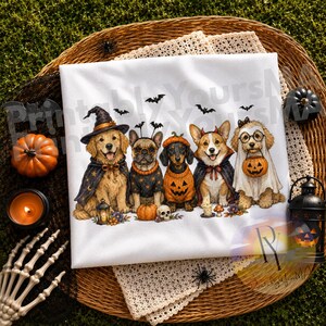 May include: A white t-shirt featuring a graphic of five dogs dressed in Halloween costumes, including a witch, a pumpkin, and a ghost, surrounded by small bats, with the text PrintablyYoursM printed faintly across the top of the fabric.