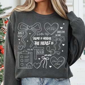 May include: Dark gray sweatshirt with white graphic elements related to real estate. The design includes bows, hearts, a key, a house, and the phrases "Just Sold," "Dream Home," and "Home is Where the Heart Is." The sweatshirt is a casual style.