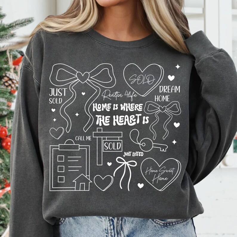 May include: Dark gray sweatshirt with white graphic elements related to real estate. The design includes bows, hearts, a key, a house, and the phrases "Just Sold," "Dream Home," and "Home is Where the Heart Is." The sweatshirt is a casual style.