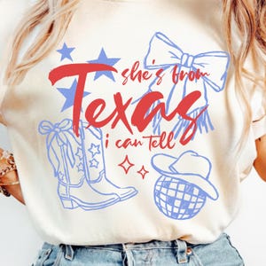 May include: Cream-colored t-shirt with a red and blue graphic design. The design includes the word "Texas" in red script, blue stars, and illustrations of a cowboy hat, boots, and a bow. The text "she's from" and "I can tell" are also included.