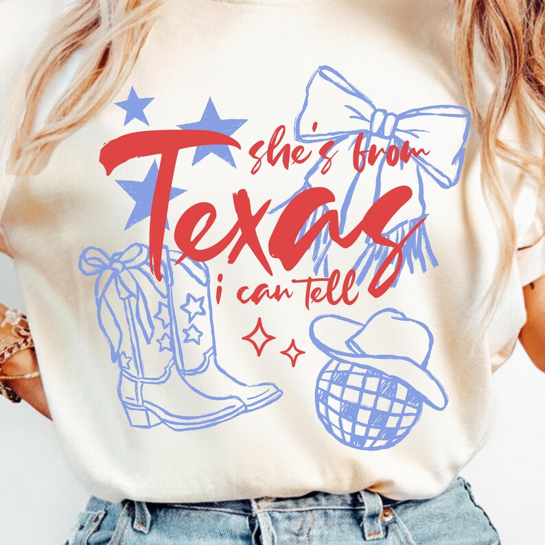 May include: Cream-colored t-shirt with a red and blue graphic design. The design includes the word "Texas" in red script, blue stars, and illustrations of a cowboy hat, boots, and a bow. The text "she's from" and "I can tell" are also included.