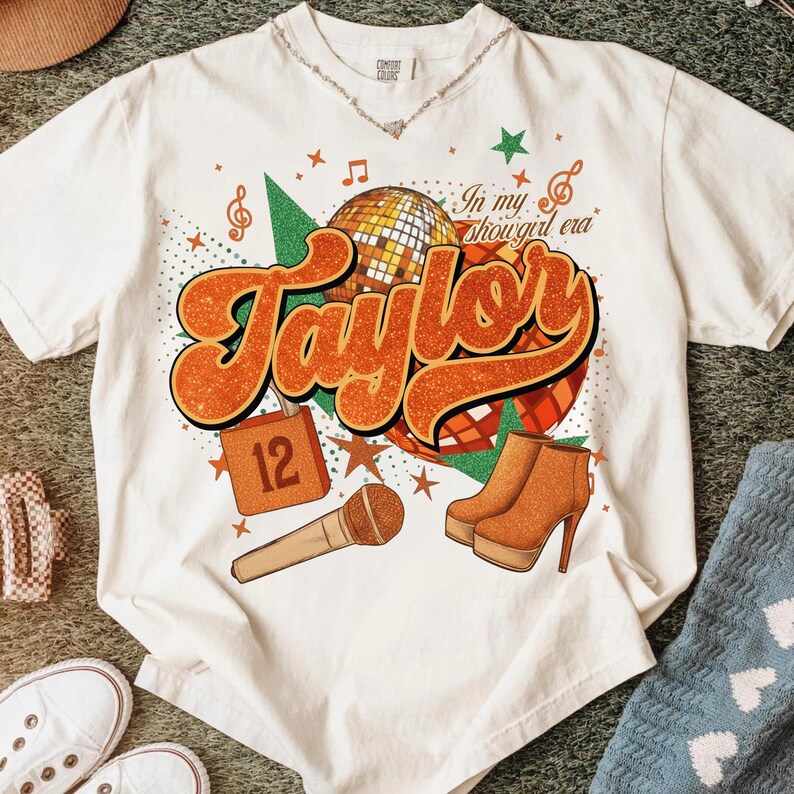 K&ouml;nnte beinhalten: Cremefarbenes T-Shirt mit Retro-Design mit dem Namen "Taylor" in Orange, einer Discokugel und einem Mikrofon. Das Shirt enth&auml;lt auch den Text "In my showgirl era" und Bilder von Stiefeln und Sternen.