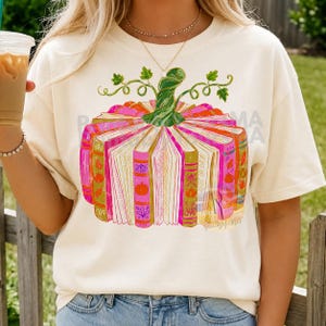 May include: A cream-coloured t-shirt featuring a graphic of a pumpkin constructed from upright books. The books are shades of pink, orange, and gold with pumpkin illustrations on the spines. A green stem sits atop the book pumpkin design.